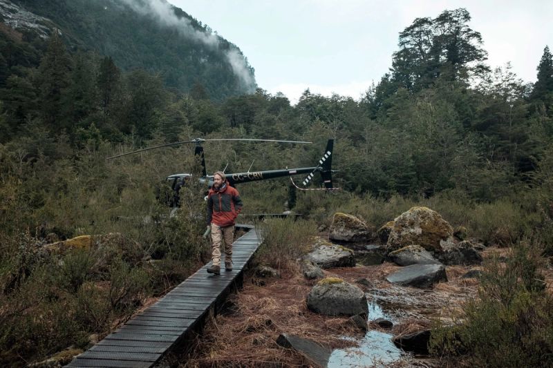 Hiker walks on wooden boardwalk near helicopter in lush Patagonian forest landscape.