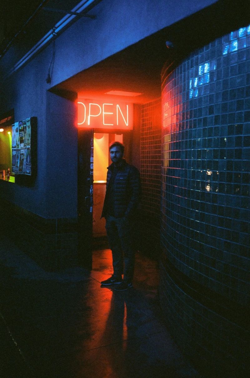 Man stands in doorway under red neon OPEN sign, blue-tiled wall to the right