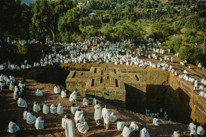 Pilgrims in white robes surround a massive stone cross church carved into the side of a hill.