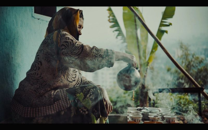 Woman in patterned dress pours tea on a misty balcony with banana trees