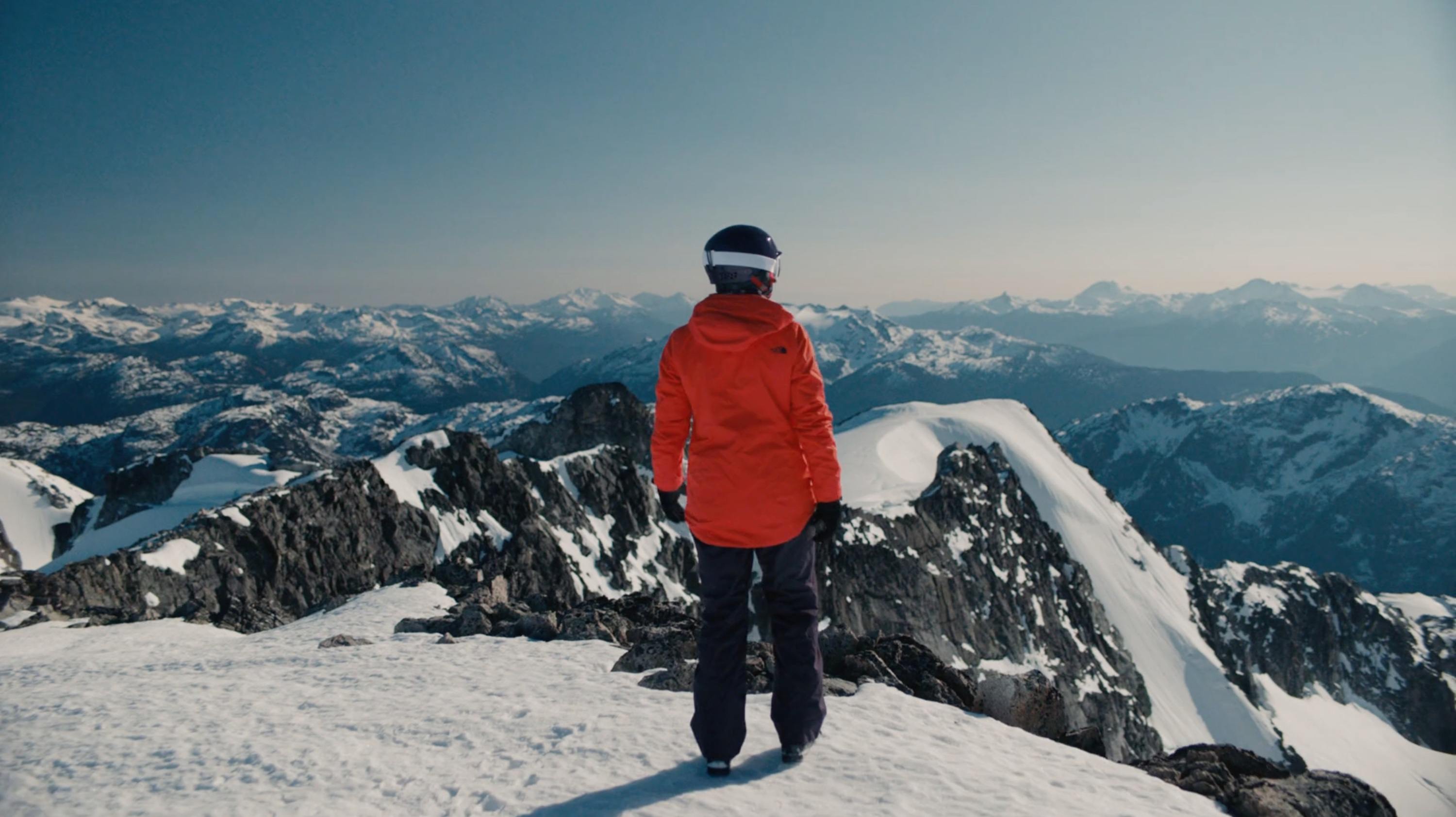 Skier in orange jacket stands on snowy mountain peak overlooking vast alpine range