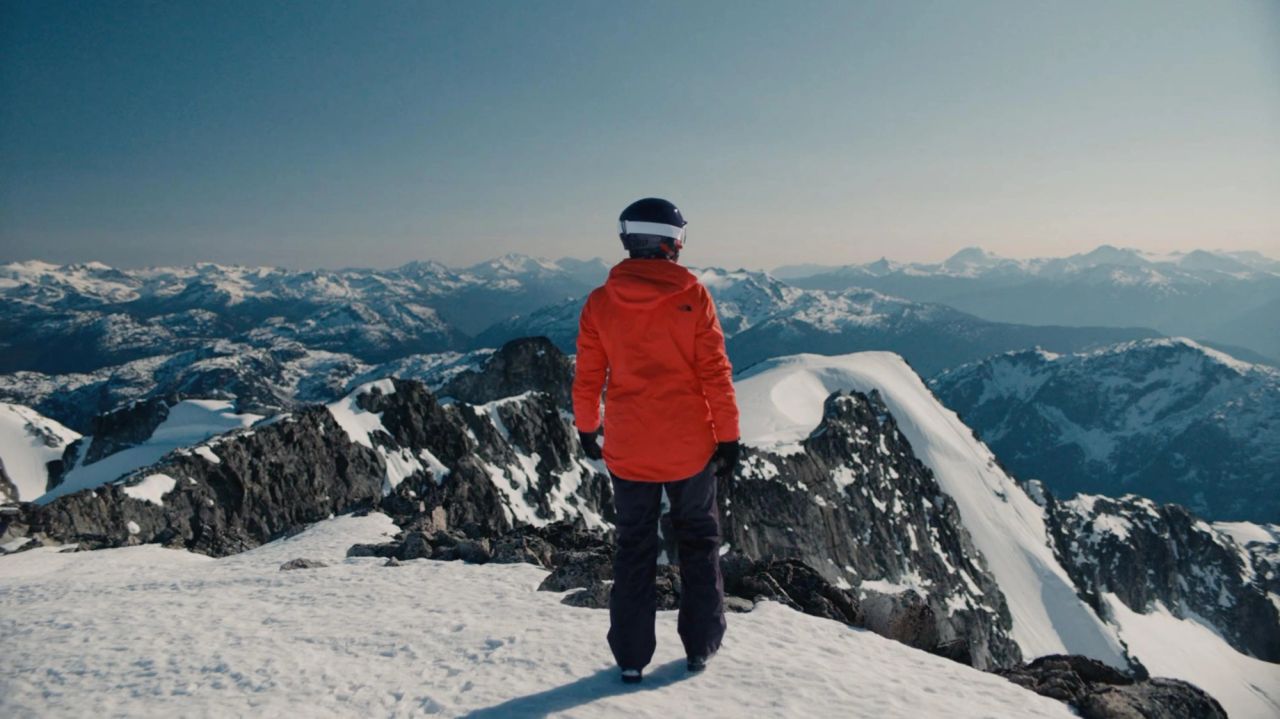 Skier in orange jacket stands on snowy mountain peak overlooking vast alpine range