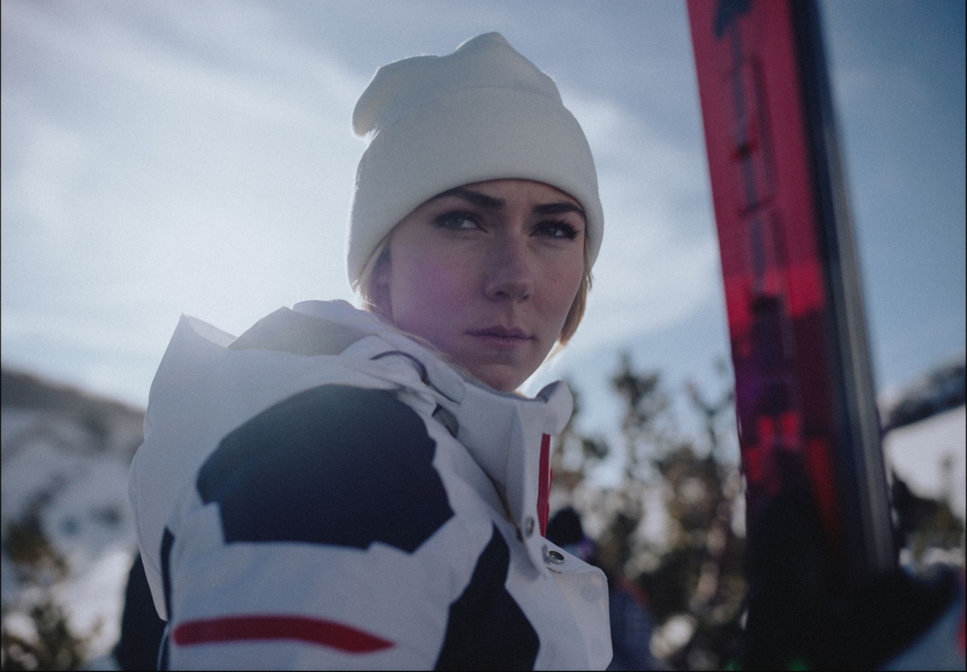 Female skier in white beanie and jacket holding red skis, snowy mountain background