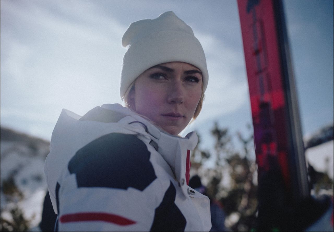 Female skier in white beanie and jacket holding red skis, snowy mountain background