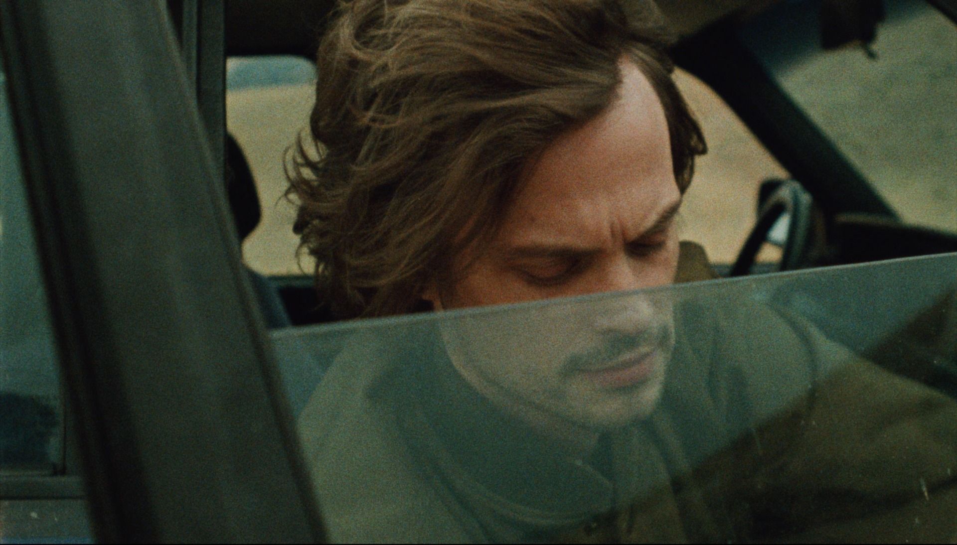 Close-up of a man with tousled hair looking down inside a car, viewed through the window.