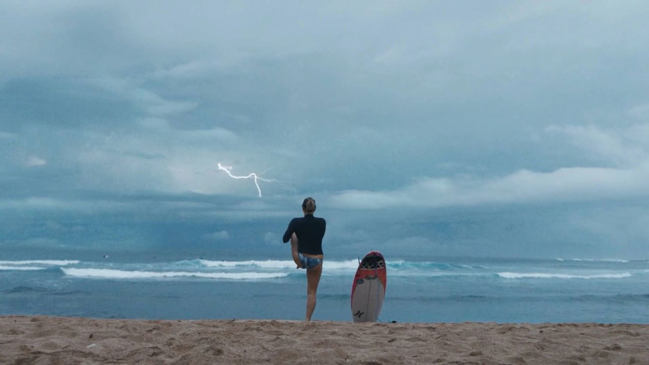 Surfer stands on beach watching lightning over ocean waves