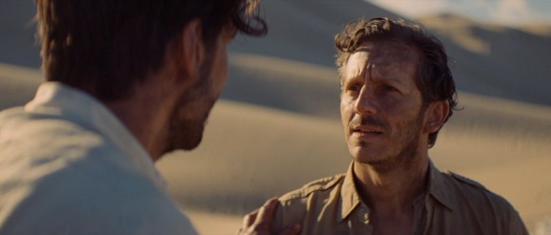 Two men talking in a desert landscape with sand dunes.