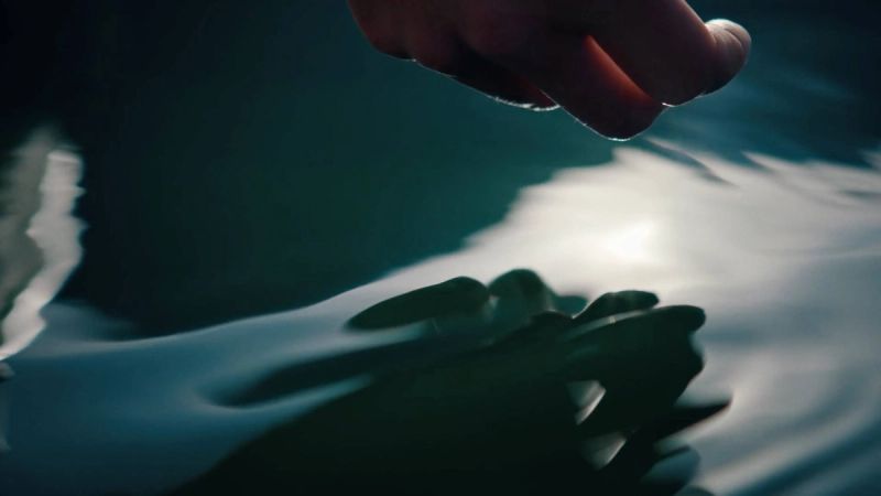 Cinematic shot of a hand reaching toward rippling water with sunlight reflecting on the surface.