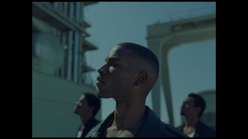 Giveon in profile, looking upward with two men behind him on a sunlit industrial structure