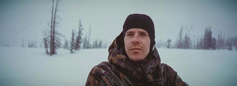 Cinematographer portrait in camouflage jacket and beanie standing in a snowy field with trees.