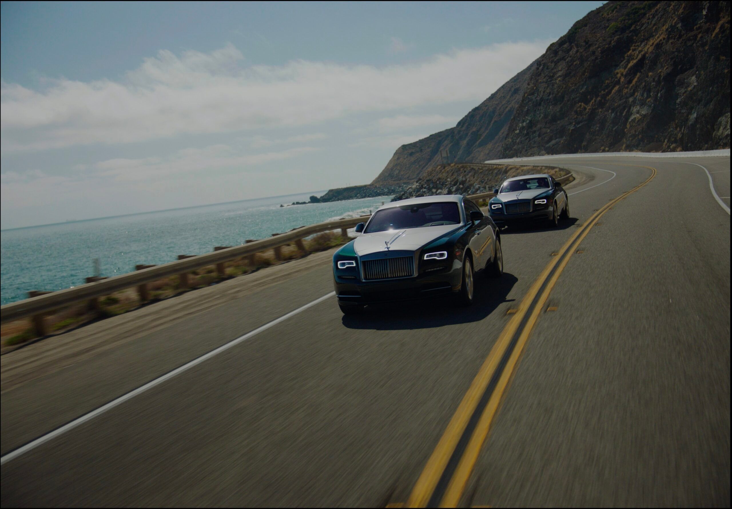 Two Rolls Royce vehicles driving along a scenic coastal highway.