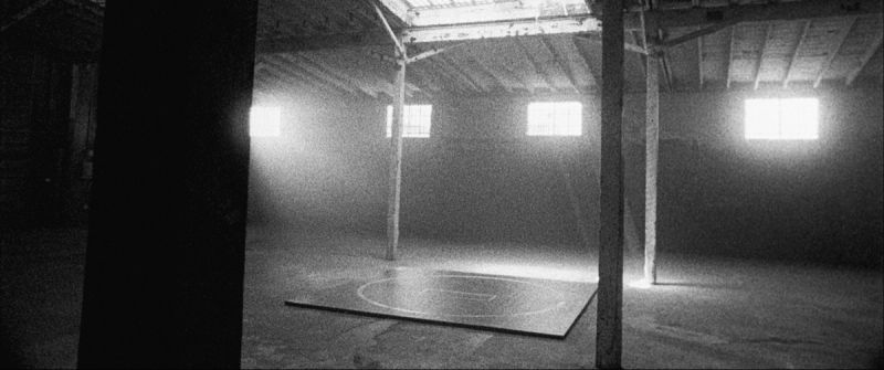 Black and white cinematography of a wrestling mat in a dimly lit industrial gym space with light streaming through windows.