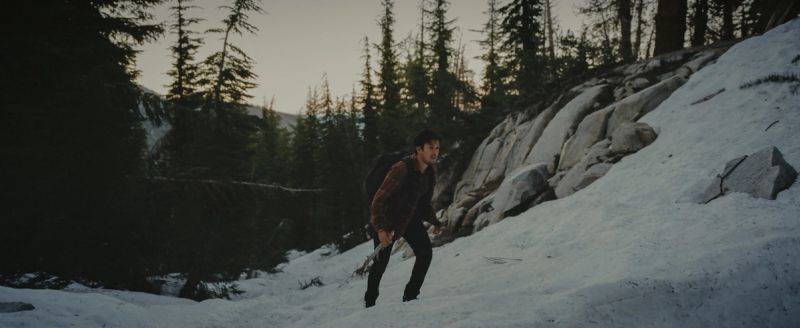 A man hiking up a snowy mountain slope surrounded by pine trees at dusk.