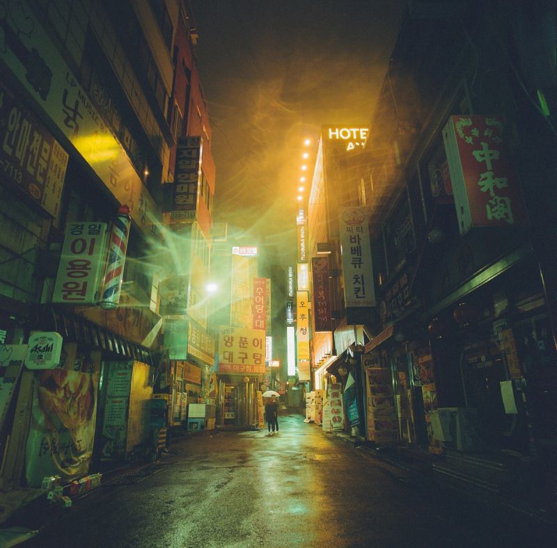 Atmospheric film photography of a wet Seoul street at night with glowing neon signs and lens flare.