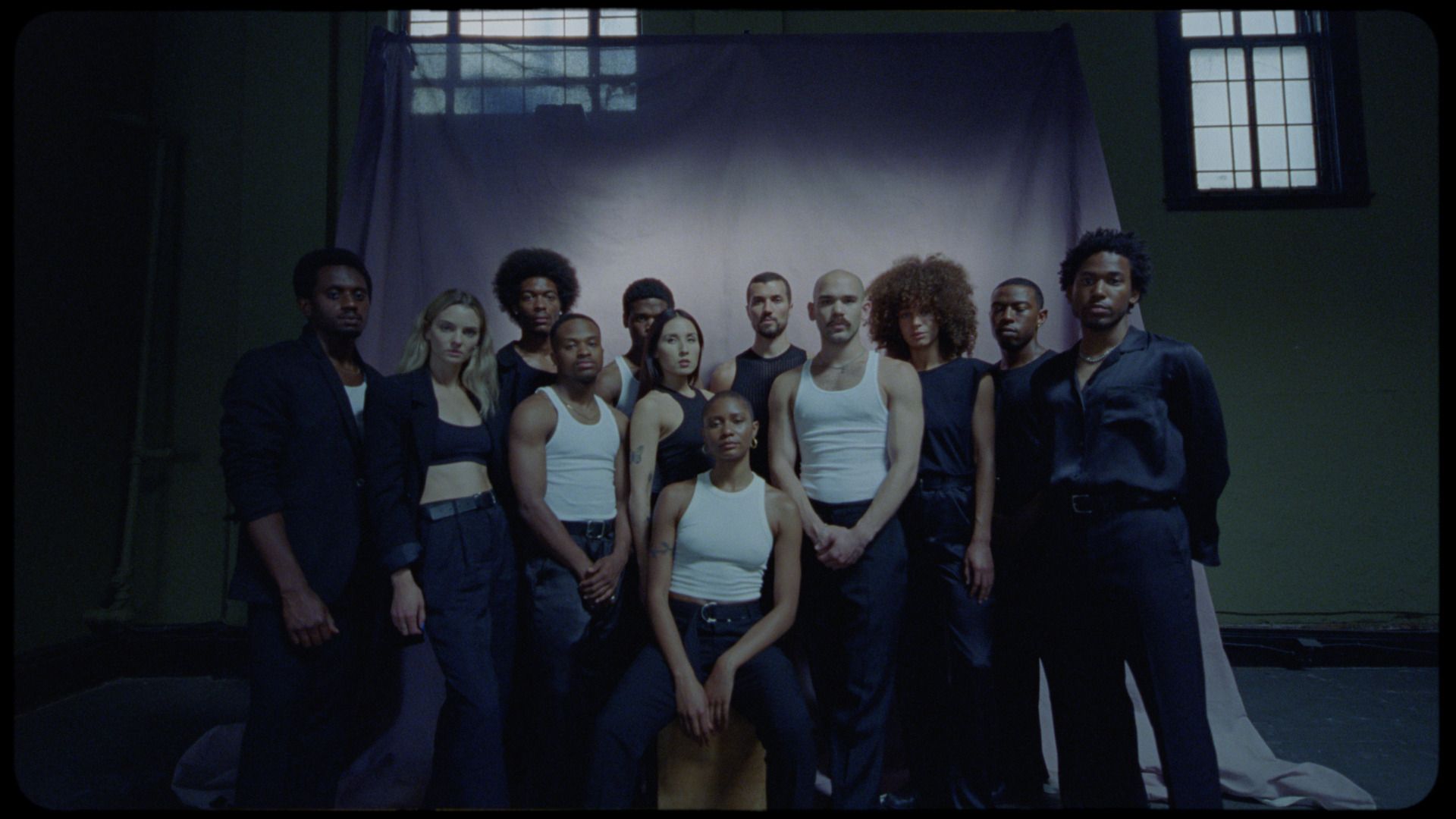 Group of diverse performers in white tank tops and dark clothing posing together on a dimly lit stage with purple backdrop