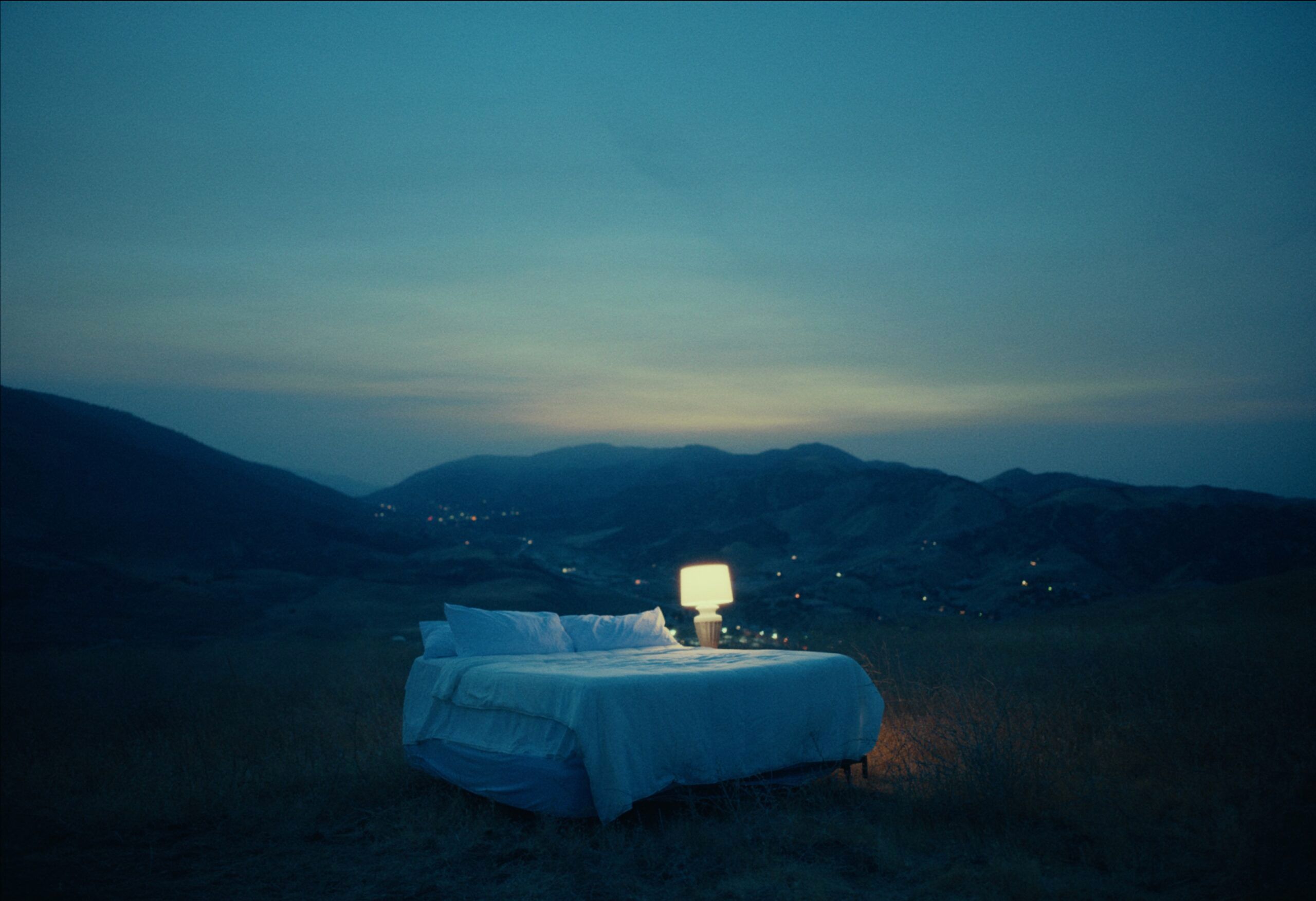 A bed with a glowing lamp sits alone in a moonlit mountain landscape at dusk.