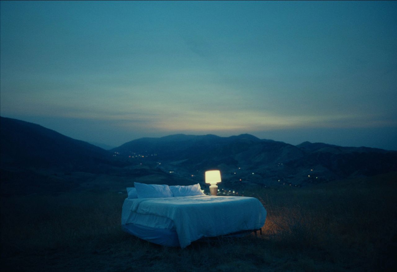 A bed with a glowing lamp sits alone in a moonlit mountain landscape at dusk.