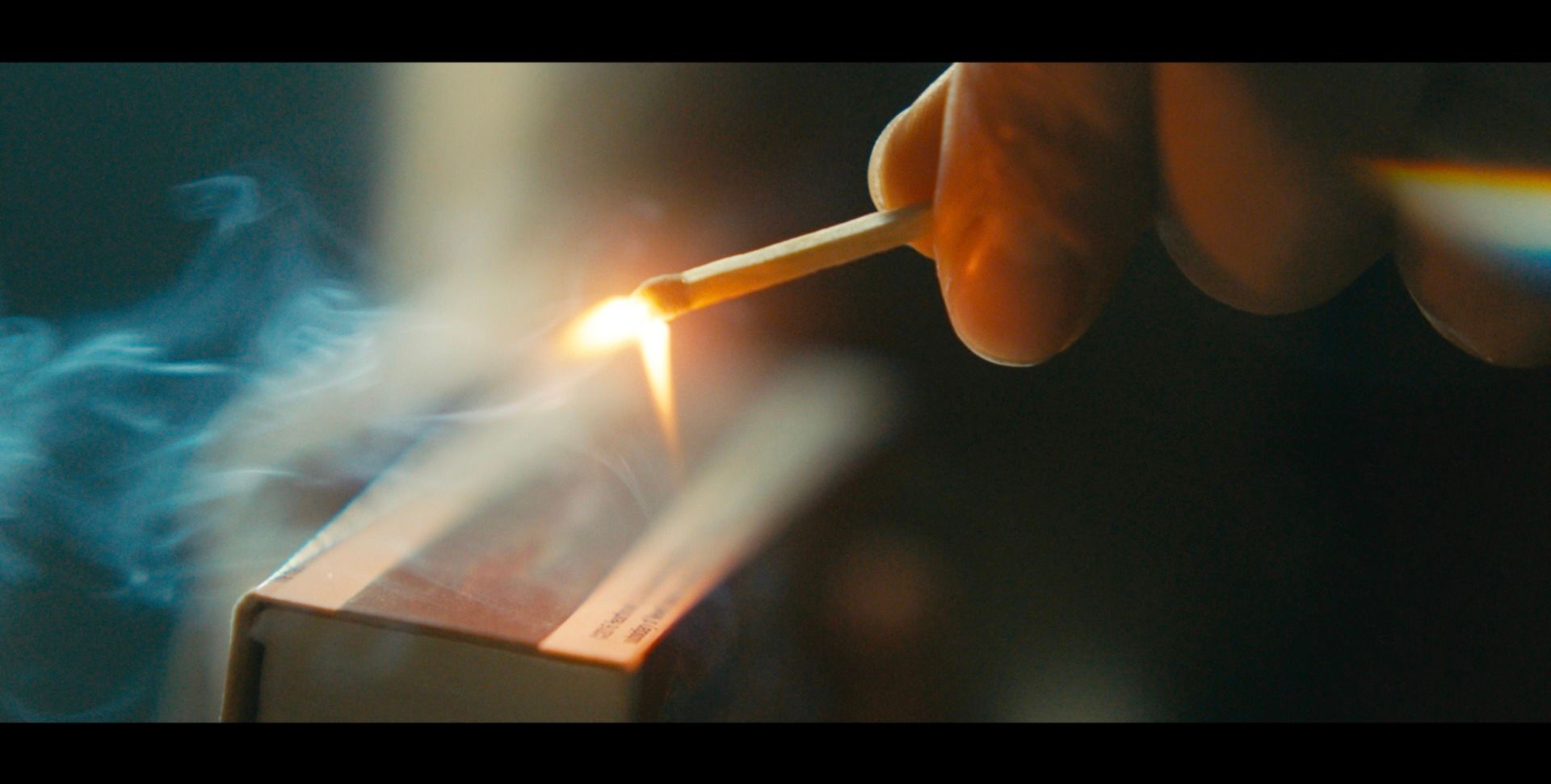 Close-up cinematography of a hand holding a lit match with smoke rising in low light.