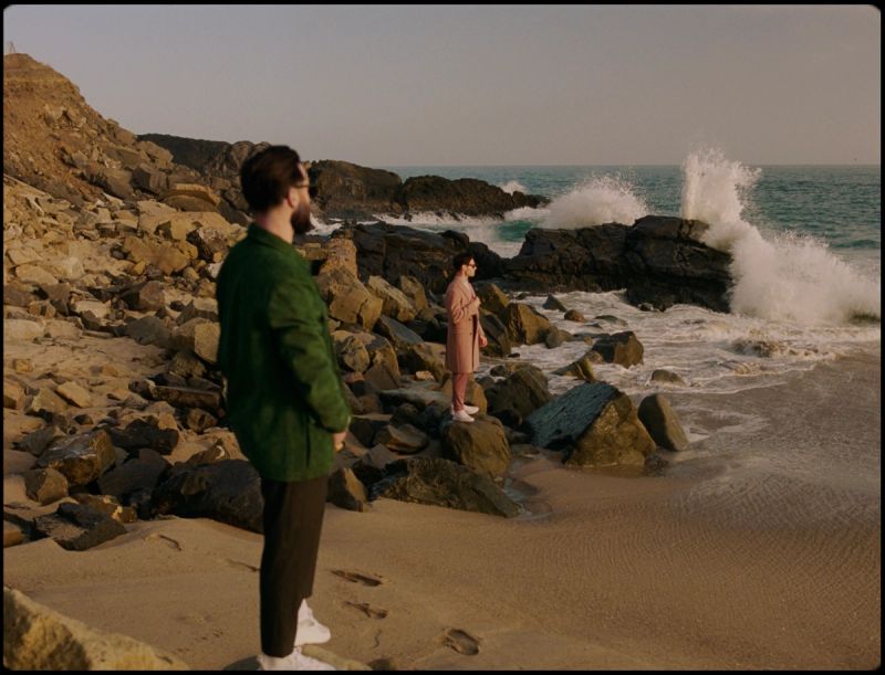 Man and woman standing on a rocky shoreline watching waves crash against dark rocks.