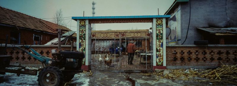 A rural courtyard with a tractor, metal gate, and brick walls under an overcast sky.