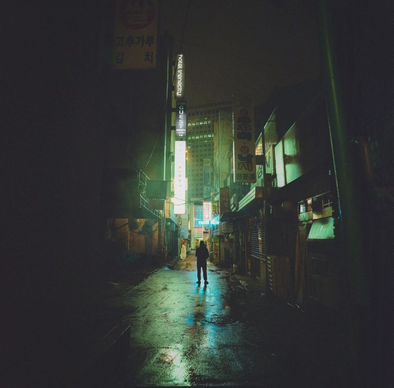 Wet night alleyway in South Korea with neon signs reflecting on pavement and lone figure.