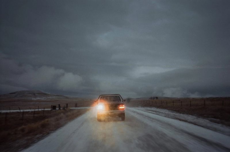 A lone car driving on a snow-covered road under an overcast sky, cinematic film photography.