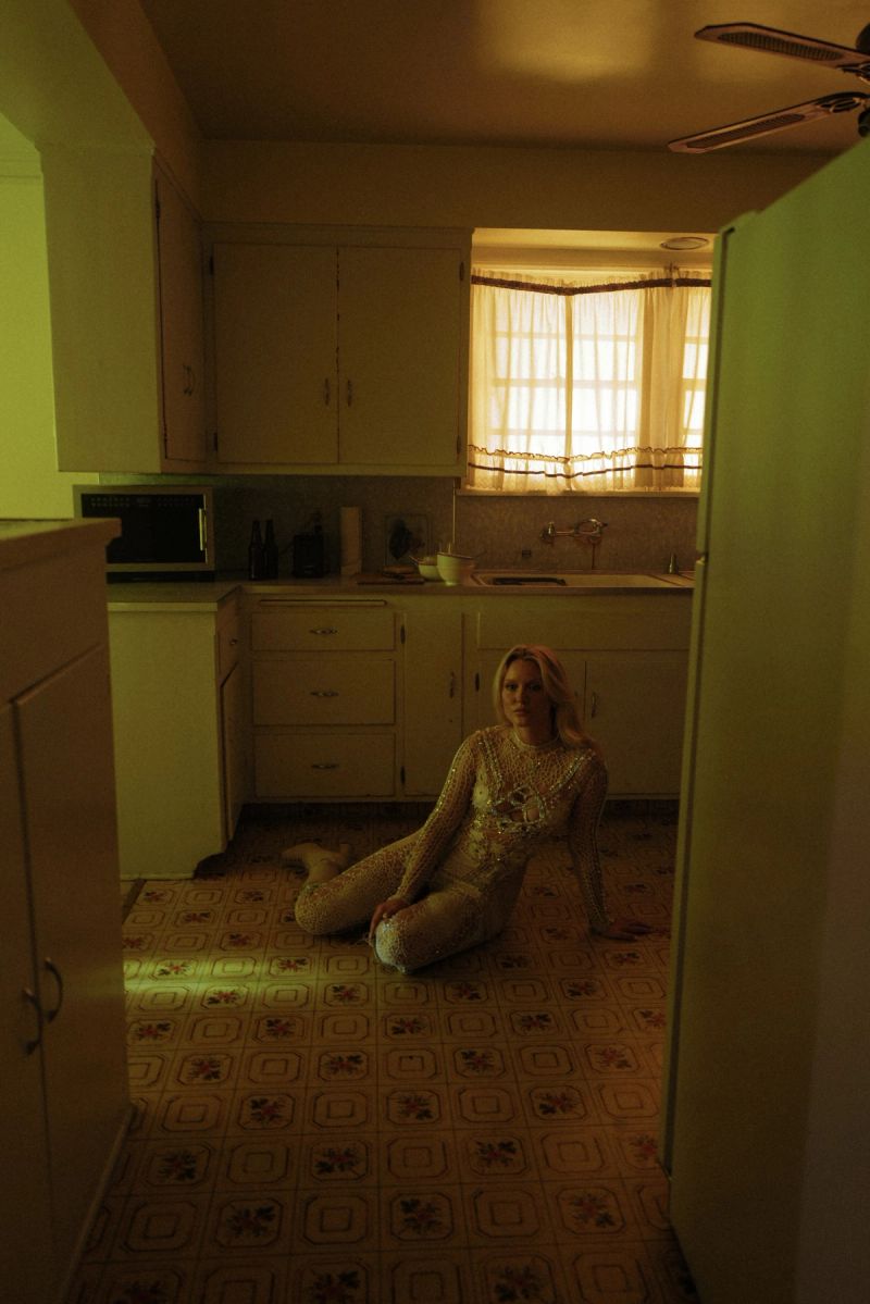 Zara Larsson in a shimmering bodysuit sitting on a vintage kitchen floor