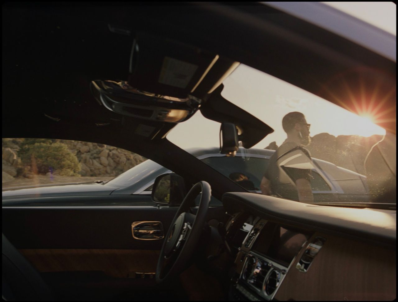 Interior view of a Rolls Royce dashboard looking out at a driver silhouetted against a bright sun flare.