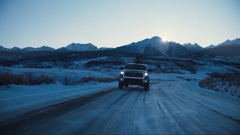 Toyota truck driving on a snowy mountain road with sun flaring over peaks.