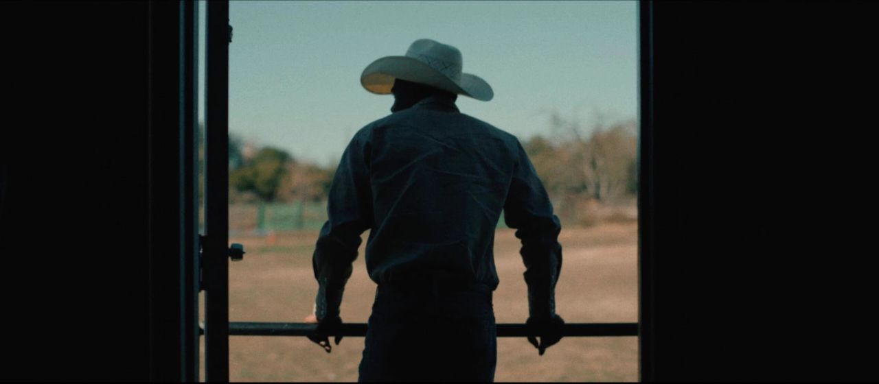 Cowboy in wide-brimmed hat stands at doorway looking out over open field