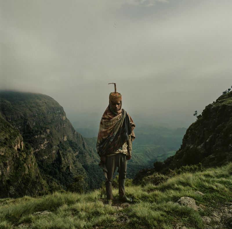 Man in traditional attire stands on a grassy cliff edge overlooking misty mountain valleys.