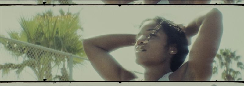 Woman looking upward outdoors with hands behind head near chain link fence and palm trees.