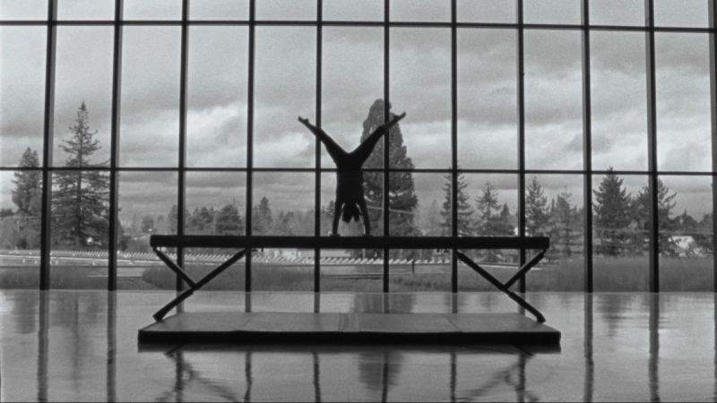 Silhouette of a gymnast doing a handstand on a balance beam in front of large windows with trees outside.