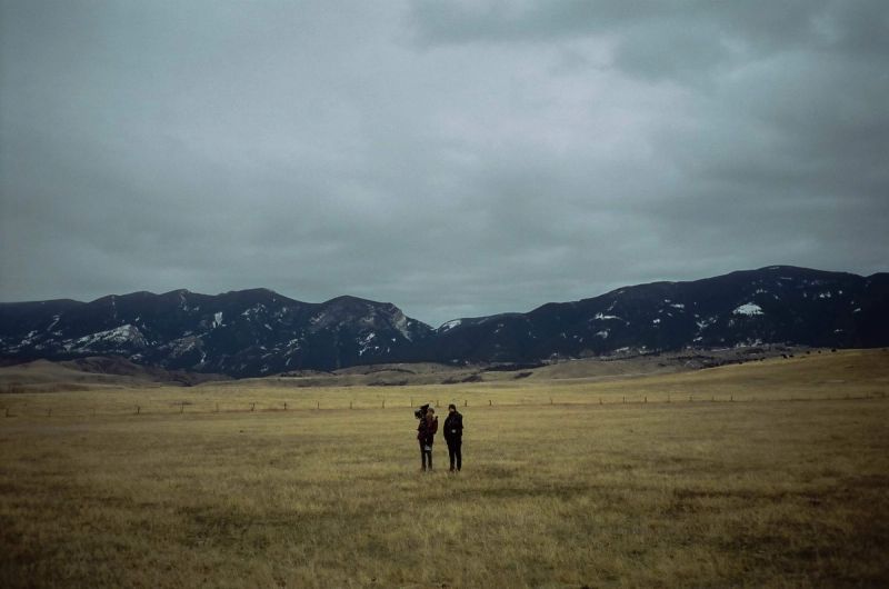 Two people stand in a vast grassy field with snow-dusted mountains under an overcast sky.