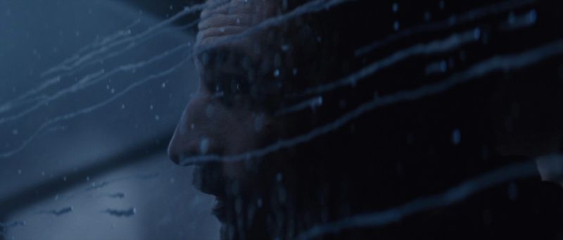 Close-up of man's face seen through rain-streaked glass in dim blue light.
