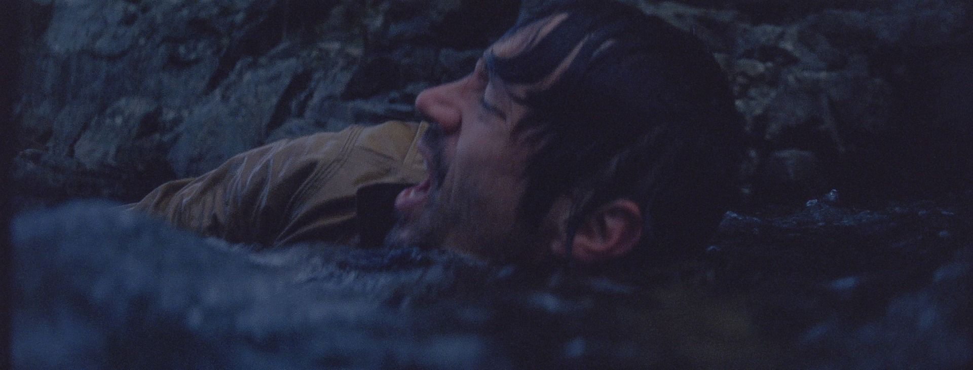 Man lying in dark rocky terrain, mouth open in distress