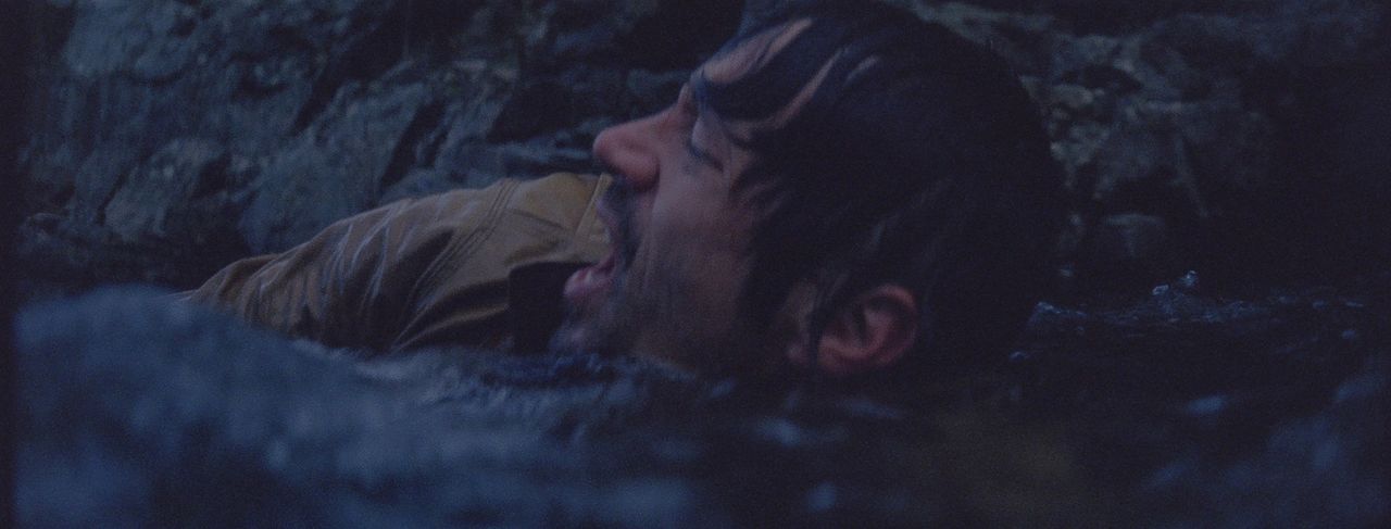 Man lying in dark rocky terrain, mouth open in distress