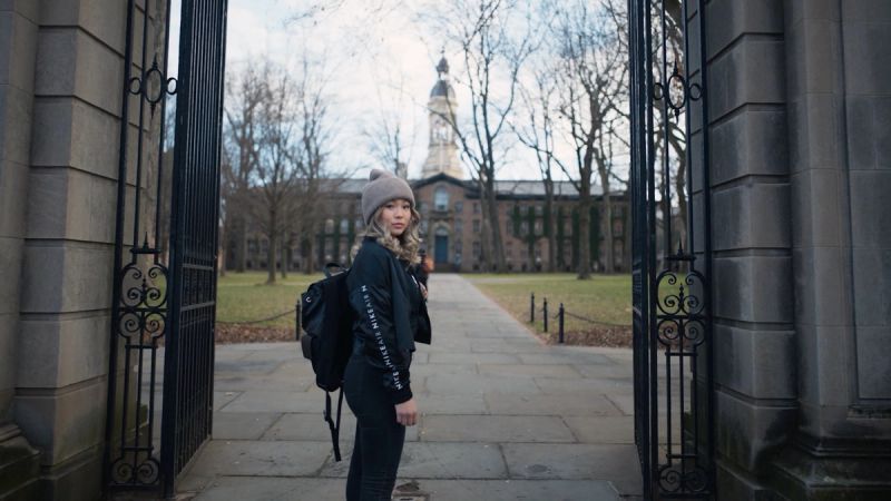 Chloe Kim stands in an open gate looking back over her shoulder, wearing a beanie and backpack on a campus path.