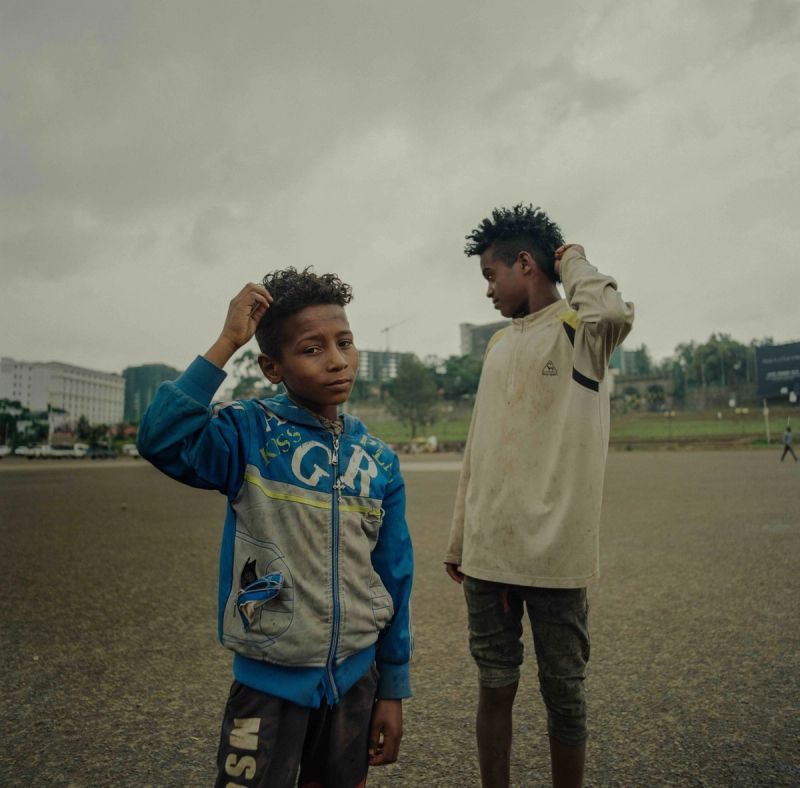 Two boys in Ethiopia stand on an open field under overcast skies, adjusting their hair.