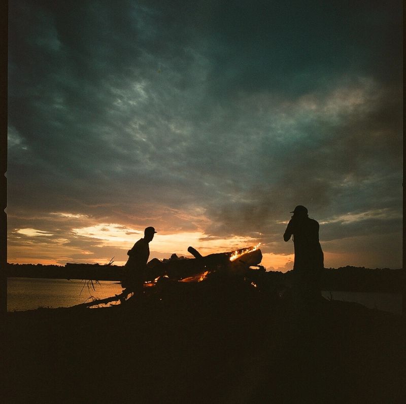 Silhouetted figures tending a campfire by the water at dusk with dramatic sunset clouds.