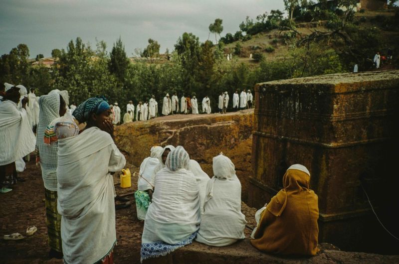 Women in traditional white robes gather near a rock-hewn structure during an outdoor ceremony.