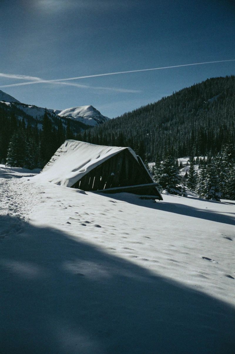 Snow-covered wooden structure in a mountain valley surrounded by pine trees.