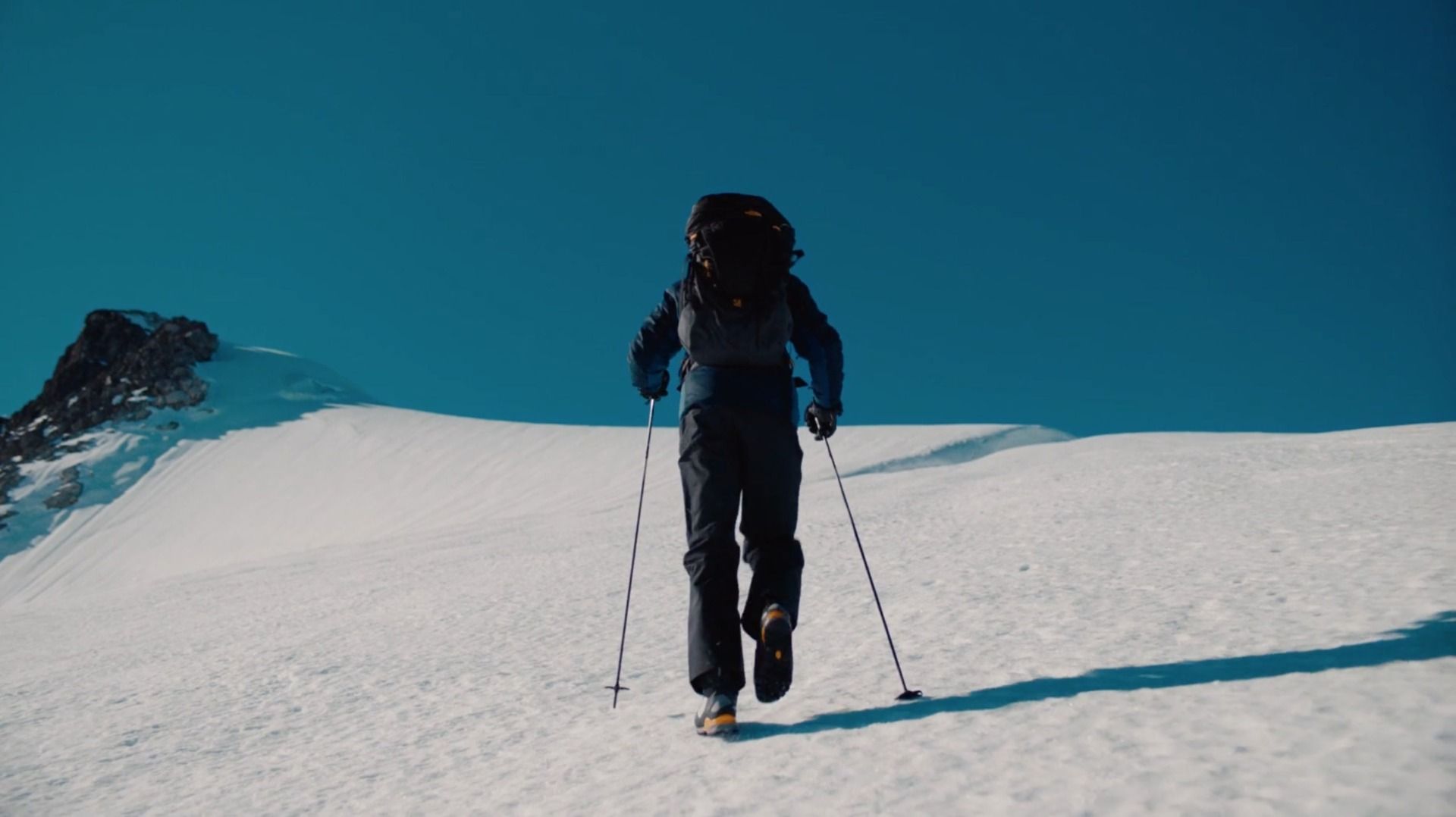 Hiker with backpack and poles ascending snowy mountain slope under clear blue sky
