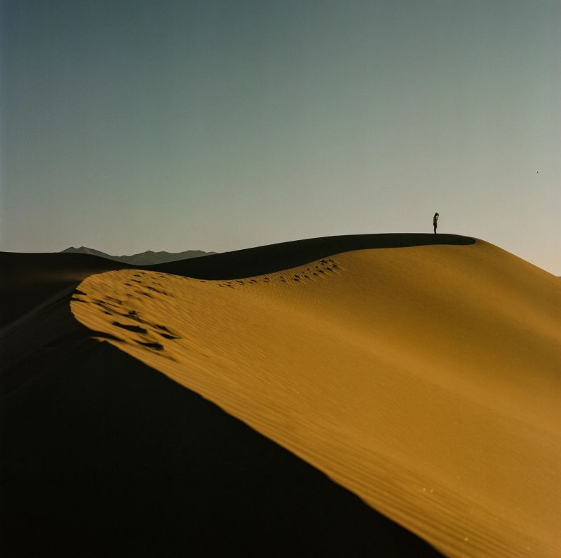 A lone silhouette stands atop a vast golden sand dune with footprints leading up the slope.
