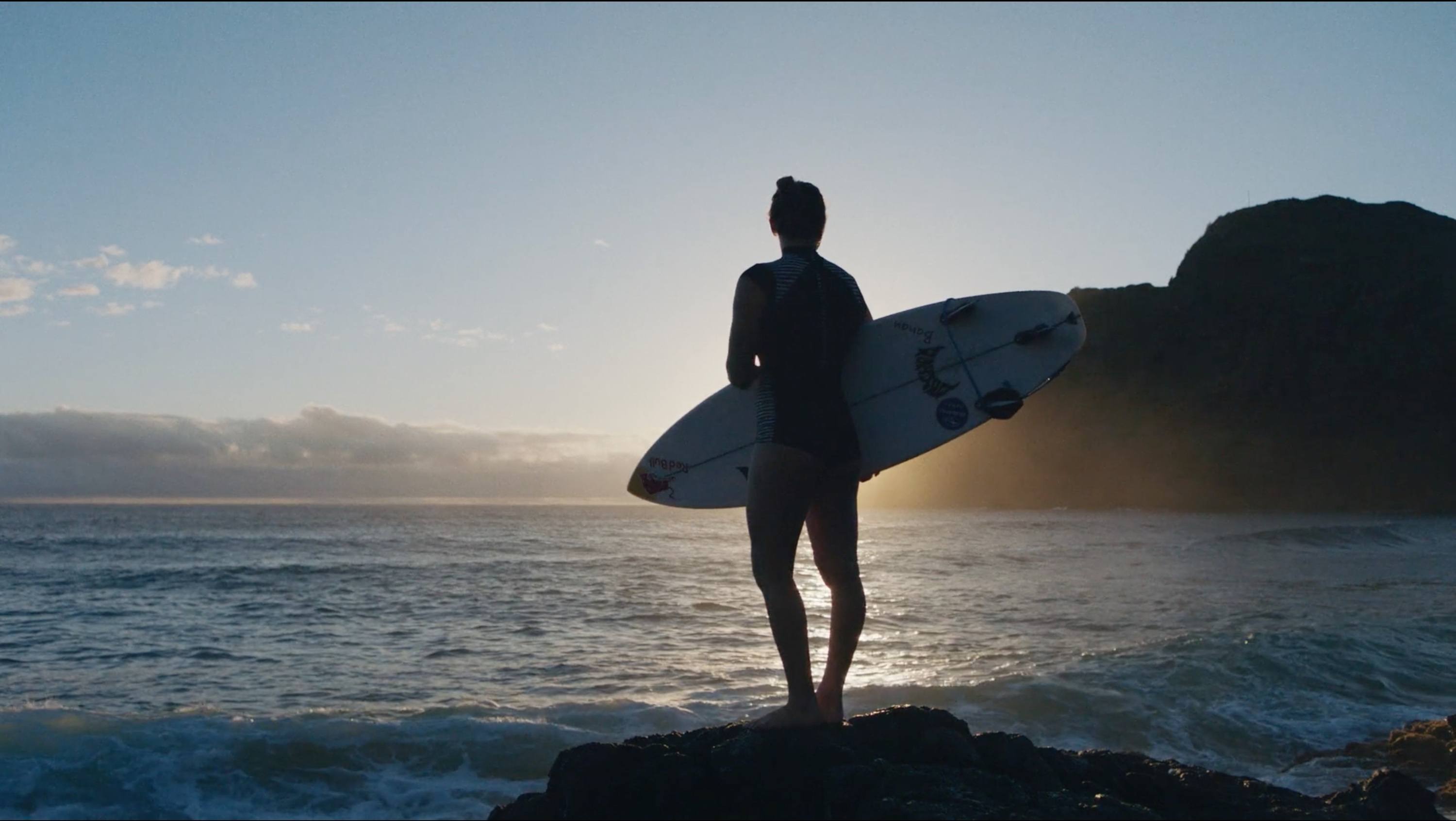 Surfer stands on rocky shore holding surfboard at sunrise over ocean