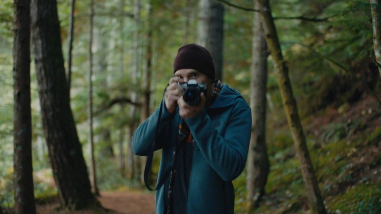 Photographer in forest holding camera, wearing beanie and blue jacket