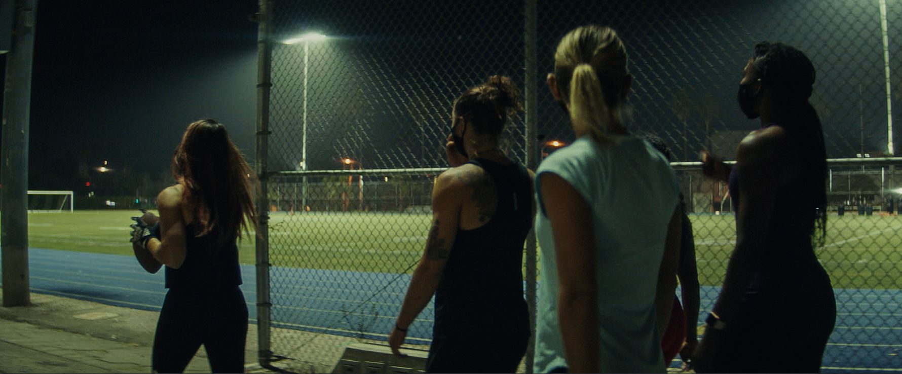 Four female athletes stand on a track at night, one holding an American football near a fence.