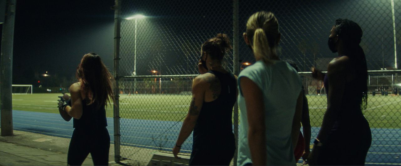 Four female athletes stand on a track at night, one holding an American football near a fence.