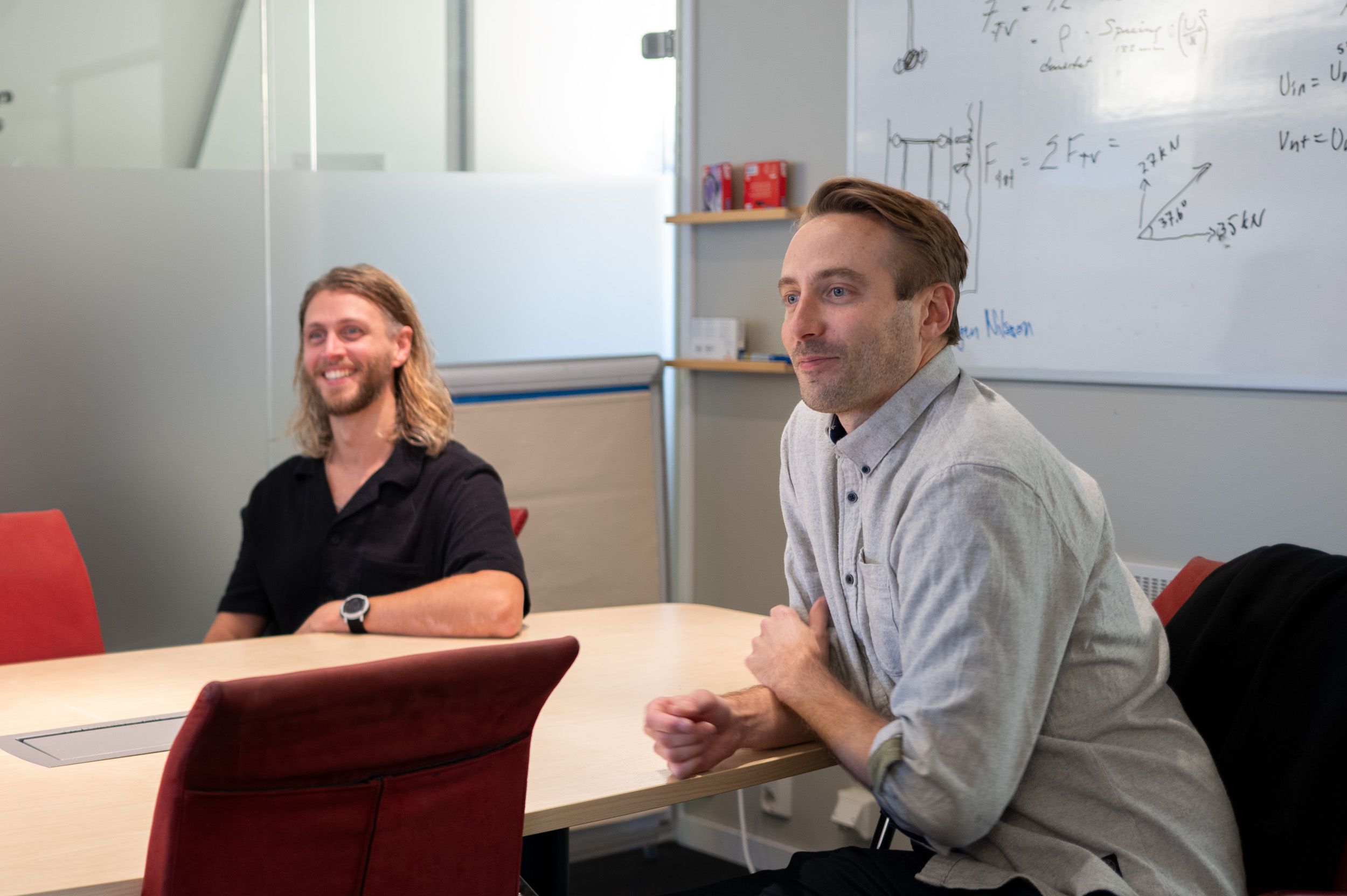 Två glada egenkonsulter under en workshop hos Topick. De sitter vid ett konferensbord och i bakgrunden syns uträkningar och skisser på en whiteboard.