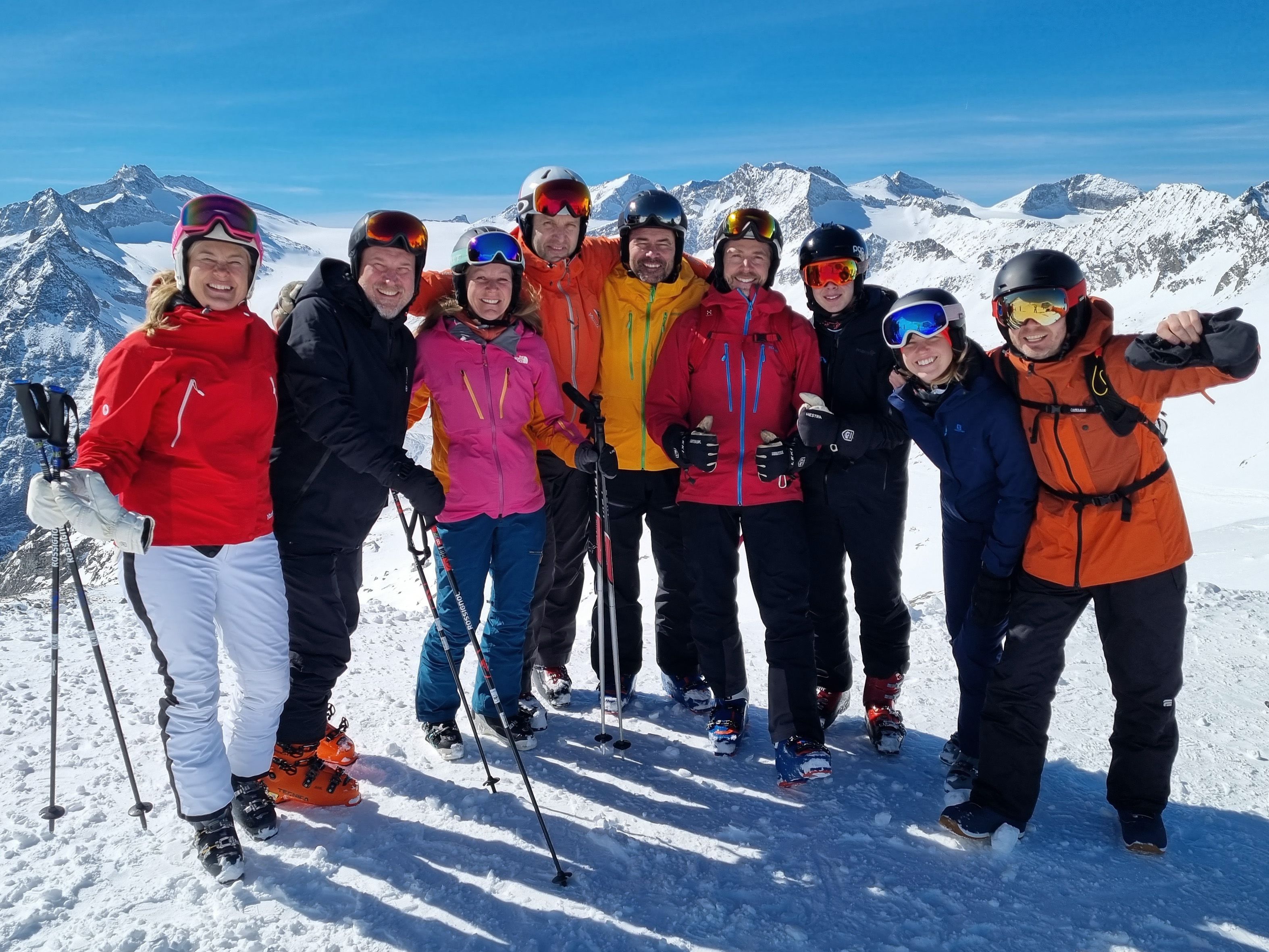 Ett gäng glada konsulter från Topick på skidsemester i alperna. Skidutrustade personer står på toppen av en skidbacke med snöiga alper i bakgrunden.